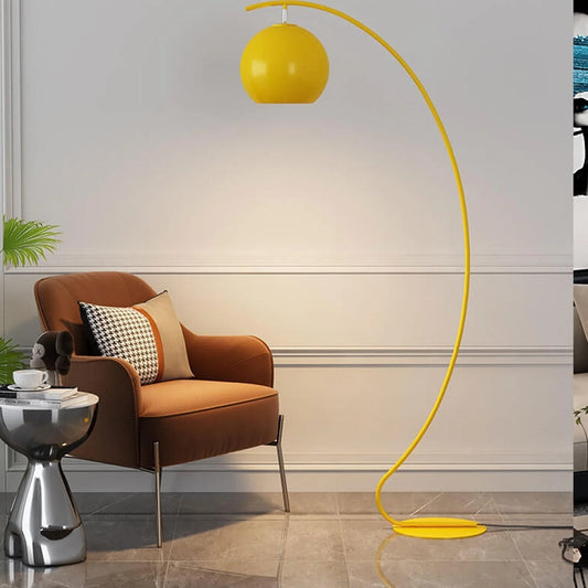 A modern yellow minimalist arc floor lamp with a spherical shade, standing next to a brown armchair in a contemporary living room.
