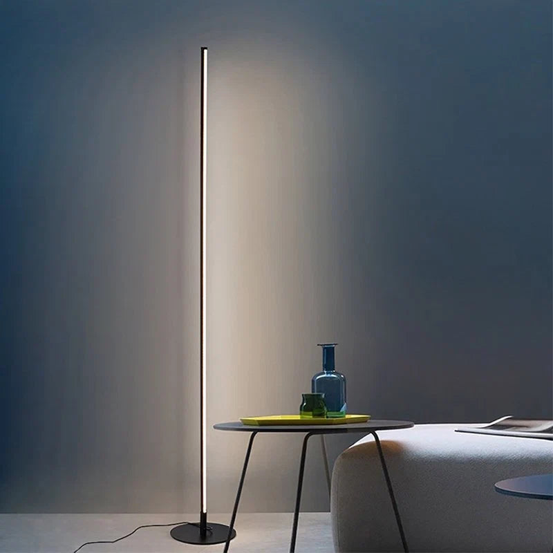 A sleek, black minimalist LED floor lamp casting a warm glow against a grey wall next to a modern side table in a contemporary living room.