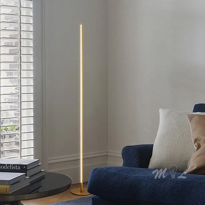 A minimalist gold LED floor lamp emitting a warm glow in a modern living room corner next to a blue armchair and window blinds.