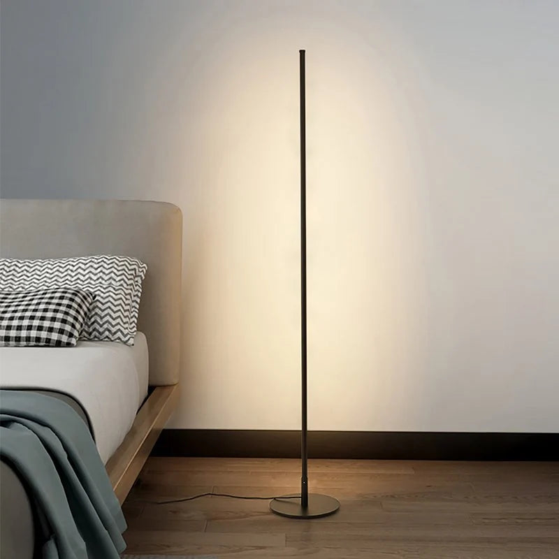A sleek, minimalist black LED floor lamp casting a warm glow against a wall, positioned next to a bed on a wooden floor in a modern bedroom.