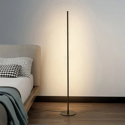 A sleek, minimalist black LED floor lamp casting a warm glow against a wall, positioned next to a bed on a wooden floor in a modern bedroom.