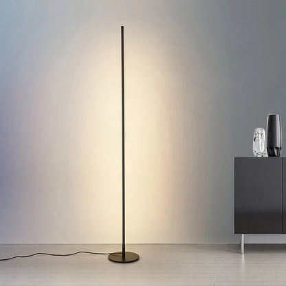 A minimalist black LED floor lamp casting a warm glow against a grey wall, standing next to a modern cabinet with decorative vases.