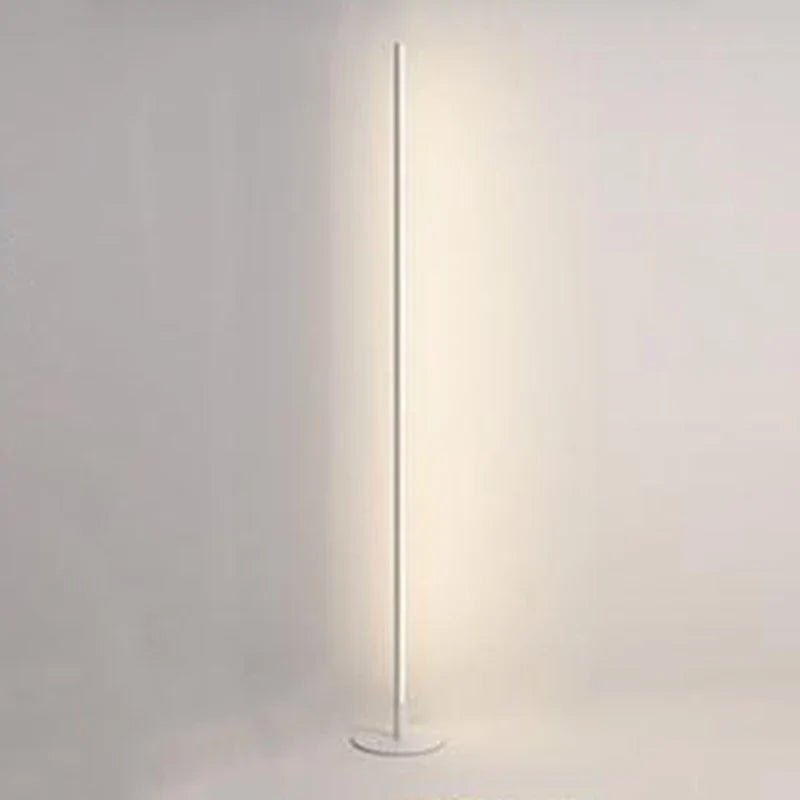 Minimalist white LED floor lamp with a slender vertical pole and circular base, emitting a soft warm glow against a plain wall.