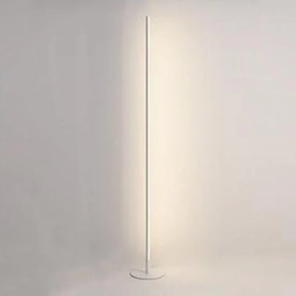 Minimalist white LED floor lamp with a slender vertical pole and circular base, emitting a soft warm glow against a plain wall.