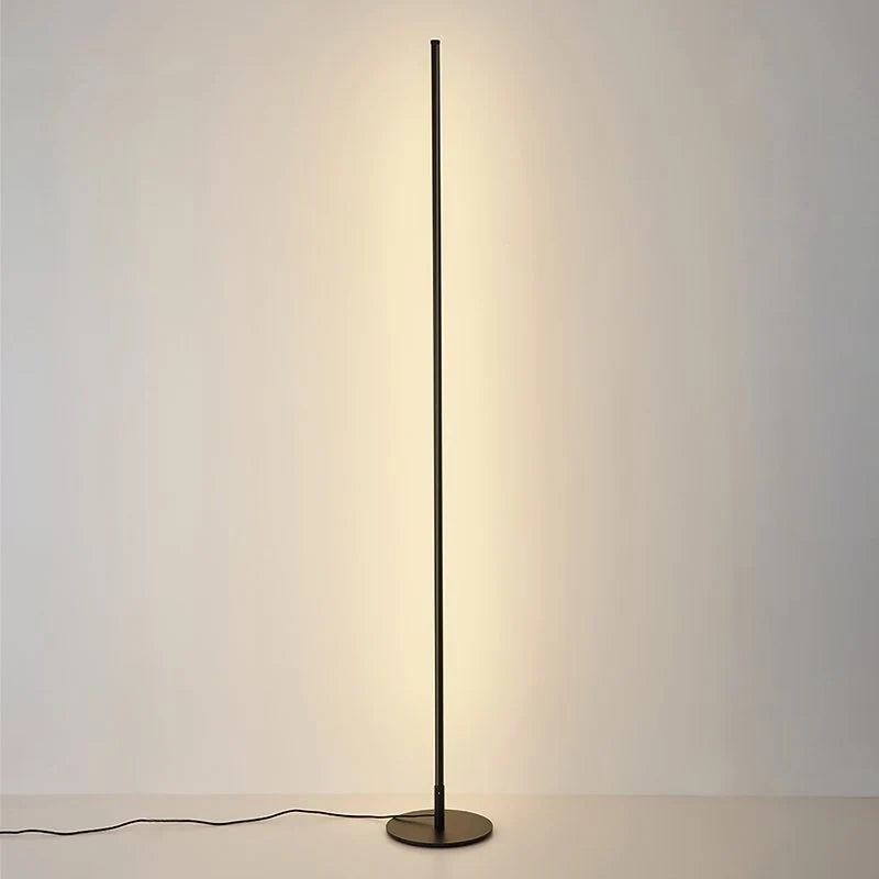 A minimalist black LED floor lamp casting a warm glow against a plain wall, featuring a slim vertical design and a circular base.