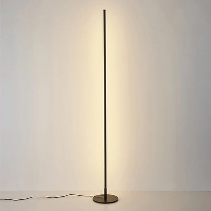 A minimalist black LED floor lamp casting a warm glow against a plain wall, featuring a slim vertical design and a circular base.