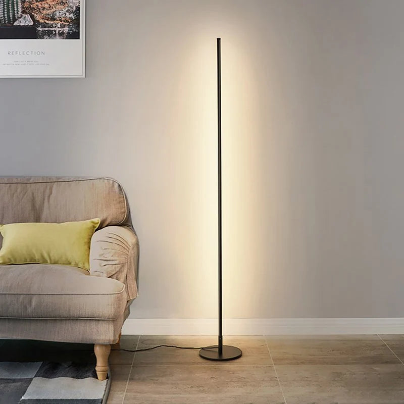 A minimalist black LED floor lamp casting a warm, soft glow against a grey wall next to a fabric armchair with a yellow cushion.