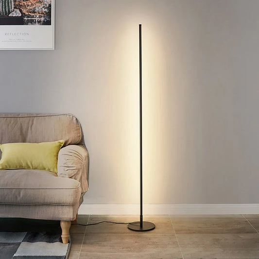 A minimalist black LED floor lamp casting a warm, soft glow against a grey wall next to a fabric armchair with a yellow cushion.