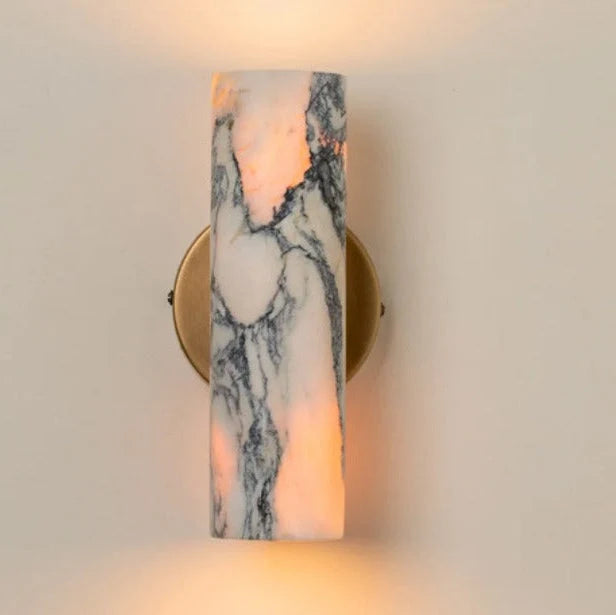 Minimalist cylindrical wall lamp featuring a white and grey marble shade mounted on a circular brass base, emitting a warm glow against a neutral wall.
