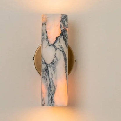 Minimalist cylindrical wall lamp featuring a white and grey marble shade mounted on a circular brass base, emitting a warm glow against a neutral wall.