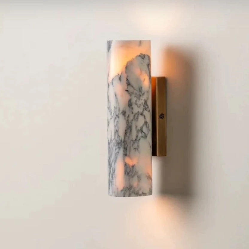 Minimalist cylindrical wall lamp featuring a white marble shade with grey veining and a brushed brass base, emitting a warm glow against a plain wall.