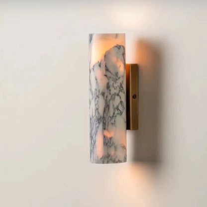 Minimalist cylindrical wall lamp featuring a white marble shade with grey veining and a brushed brass base, emitting a warm glow against a plain wall.