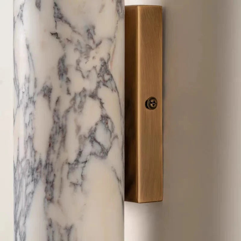 Close-up of a minimalist wall lamp featuring a cylindrical white marble shade with grey veining attached to a rectangular brushed brass mounting base.