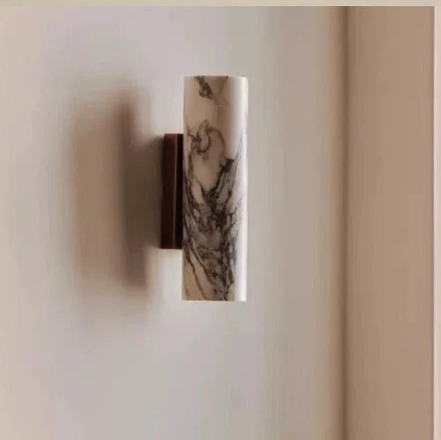 Minimalist cylindrical wall lamp featuring a white marble shade with grey veining mounted on a sleek brass base against a neutral interior wall.