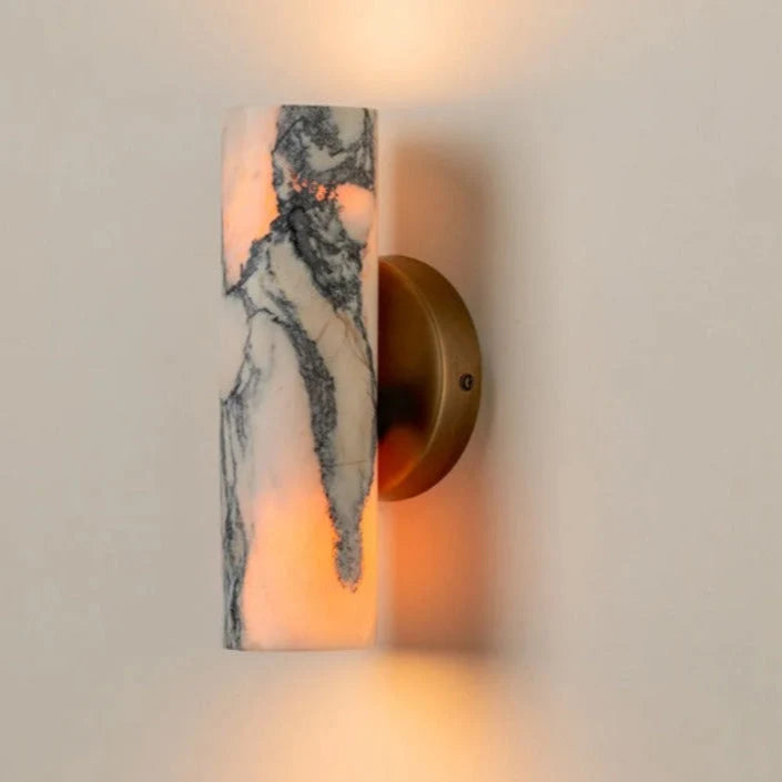 Minimalist cylindrical wall lamp featuring a white and grey marble shade mounted on a circular brass base, emitting a warm glow against a neutral wall.