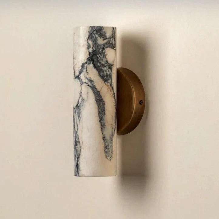 Minimalist round wall lamp featuring a cylindrical white marble shade with grey veining mounted on a circular brass base against a neutral wall.