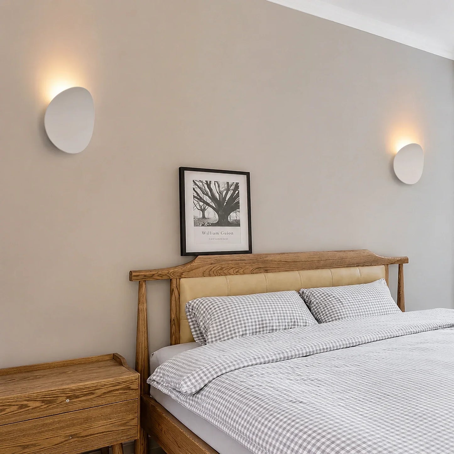 Two modern white aluminium LED wall lights providing soft up and down illumination on a neutral bedroom wall above a wooden bed frame and bedside table.