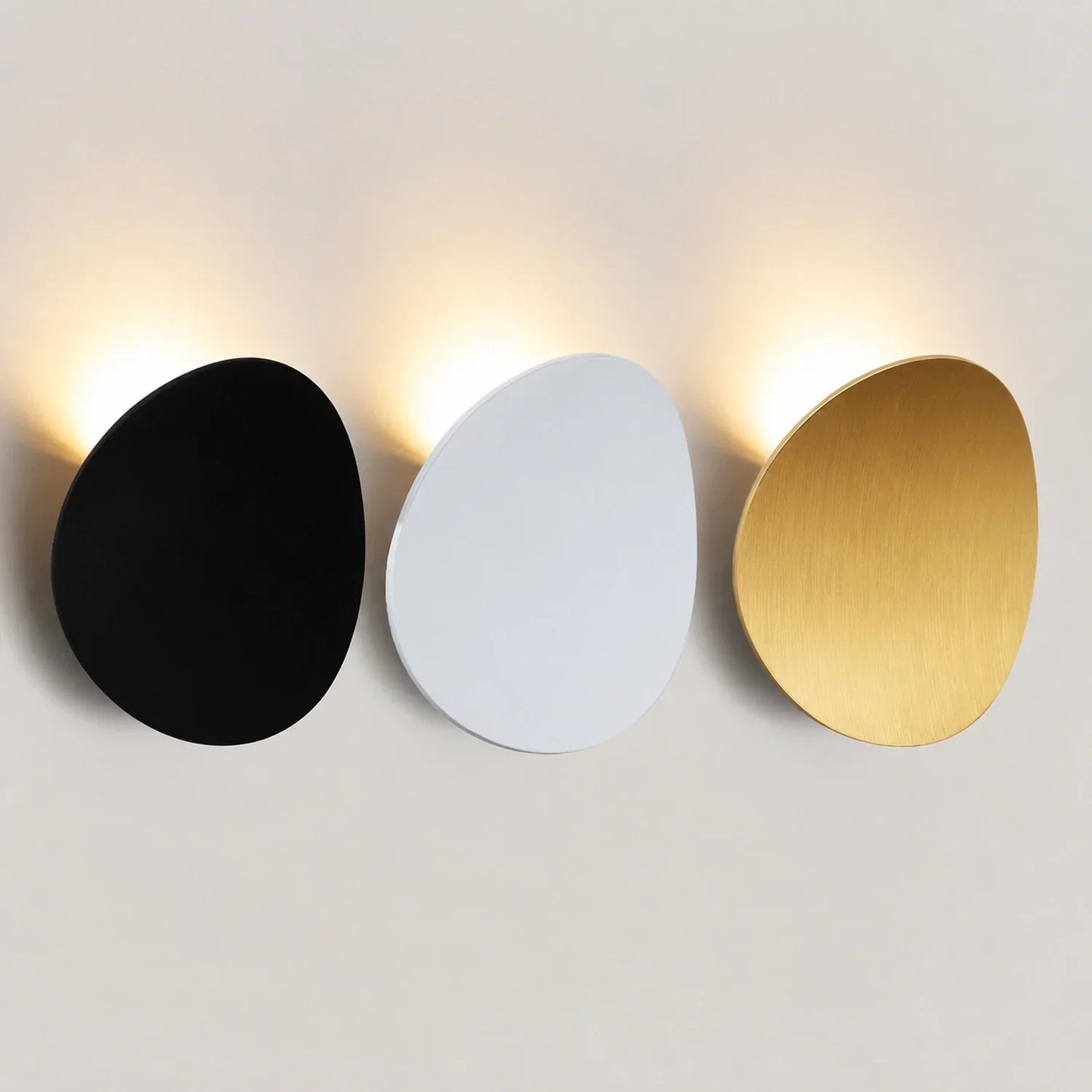 Three modern, pebble-shaped LED wall lights in black, white, and gold finishes, mounted on a wall with soft, warm up and down illumination.
