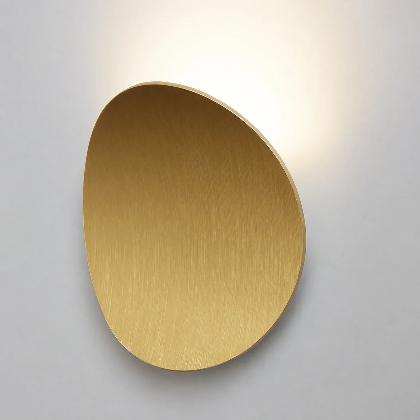 Modern brushed gold aluminium wall light with a curved, organic shape casting a soft, warm glow against a plain white wall in a contemporary interior.