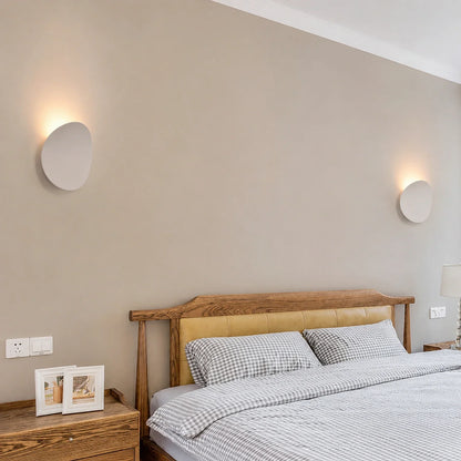 Two modern white circular LED wall lights mounted on a beige bedroom wall above a wooden bed frame, providing soft ambient up and down illumination.