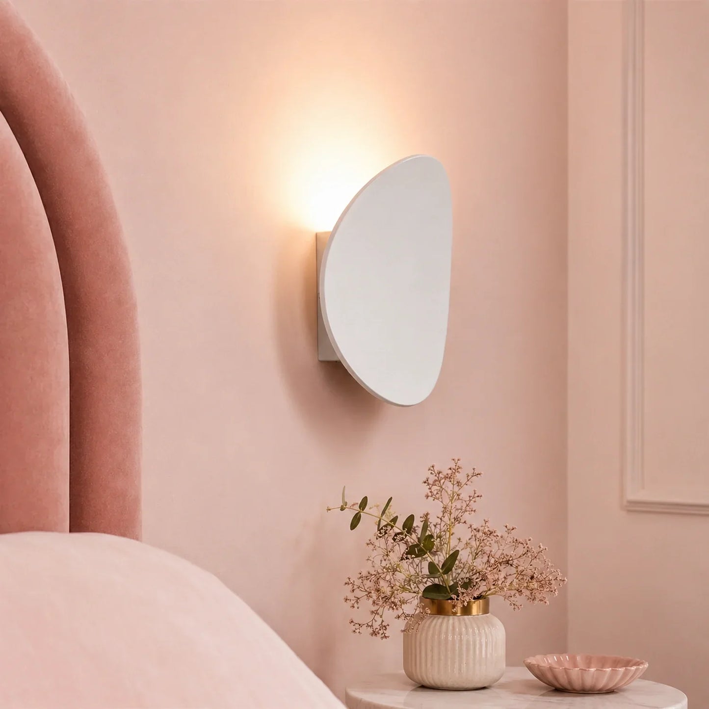 Modern white aluminium LED wall light mounted on a soft pink bedroom wall, casting a warm glow above a bedside table with a decorative vase and bowl.