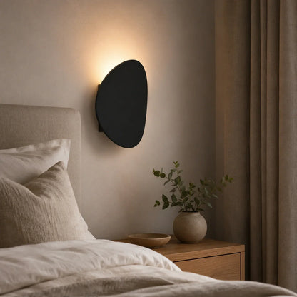 Modern black organic-shaped LED wall light mounted above a bedside table, casting a warm glow on a neutral bedroom wall with a soft, ambient effect.
