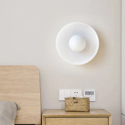 Modern Bauhaus style white flush mount ceiling light with a spherical glass shade, mounted on a neutral wall above a wooden bedside table in a bedroom.