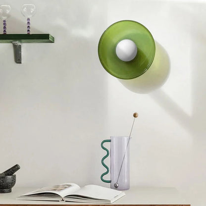 Modern Bauhaus style flush mount ceiling light in green glass with a central white bulb, mounted on a white wall above a desk with decorative items.