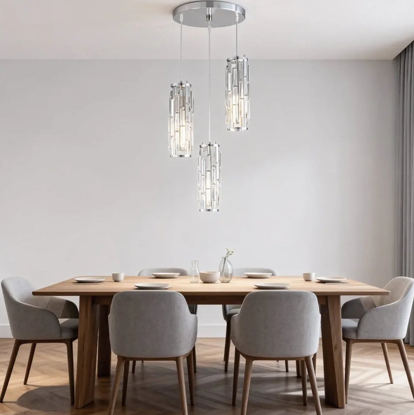A modern three-light crystal pendant fixture hanging at staggered heights over a wooden dining table with grey chairs in a bright dining room