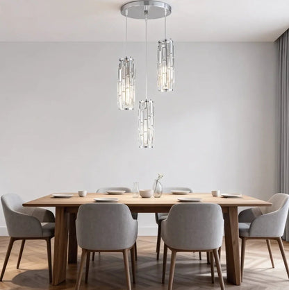 A modern three-light crystal pendant fixture hanging at staggered heights over a wooden dining table with grey chairs in a bright dining room