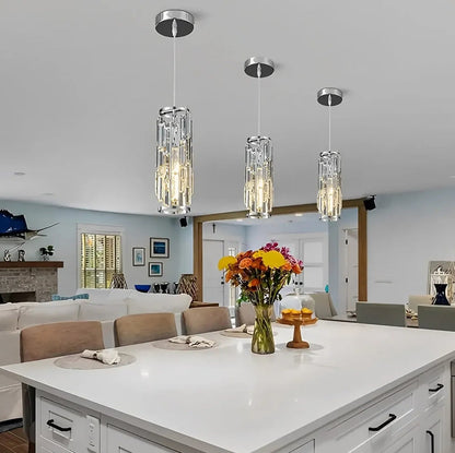 Trio of modern chrome and crystal pendant lights with a warm glow, hanging over a white kitchen island in a stylish open-plan dining area.