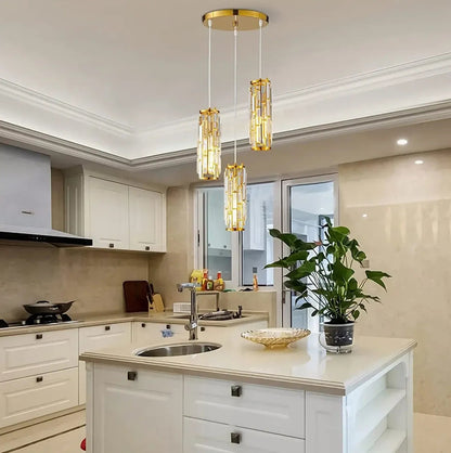 A modern three-light gold crystal pendant light fixture with a warm glow, suspended above a white kitchen island in a contemporary home.
