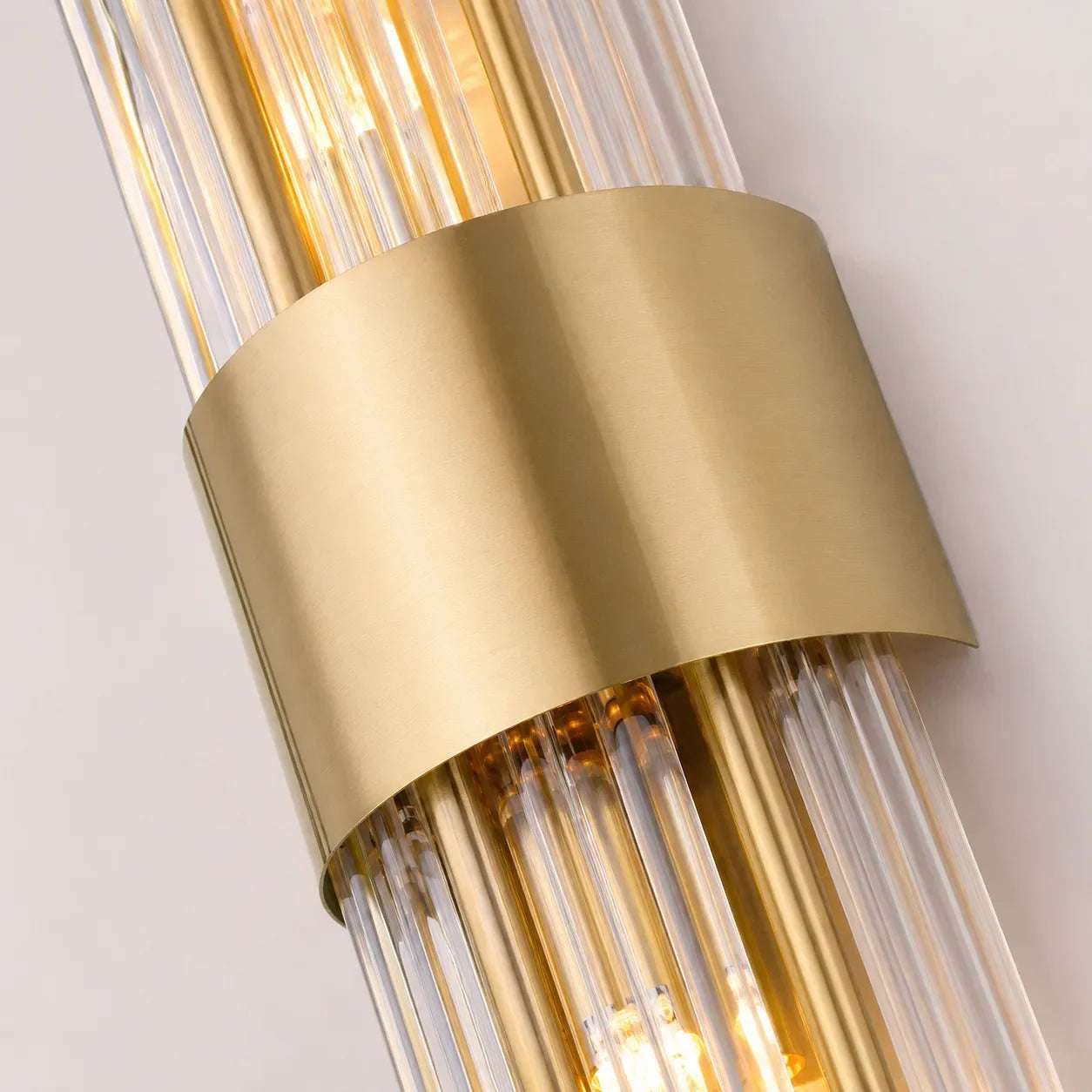 Close-up of a modern crystal wall light showing vertical glass rods encased in a brushed gold metal band, illuminated with a warm glow.