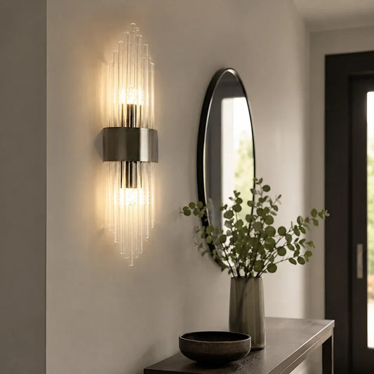 Modern crystal wall light with a black metal frame, emitting a warm white glow on a hallway wall next to a round mirror and console table.