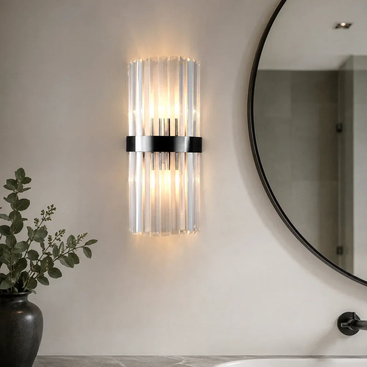 Modern 30cm wall light with a black metal frame and vertical crystal rods, emitting a warm glow on a neutral bathroom wall next to a round mirror.