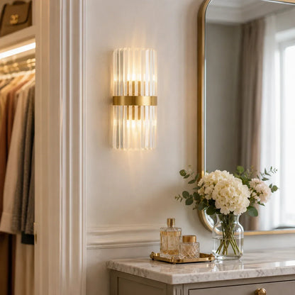 Modern crystal wall light with a gold metal frame emitting a warm white glow, mounted on a wall above a marble vanity table with a mirror.
