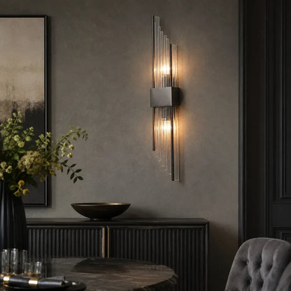 A modern black metal wall light with vertical crystal rods emitting a warm glow, mounted on a dark grey wall in a contemporary dining room.
