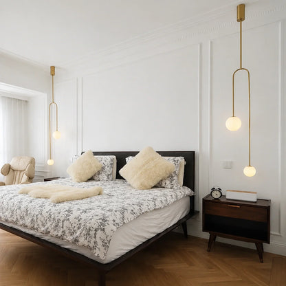 Modern double globe pendant lights with gold fixtures hanging on either side of a bed in a minimalist bedroom with white panelled walls.