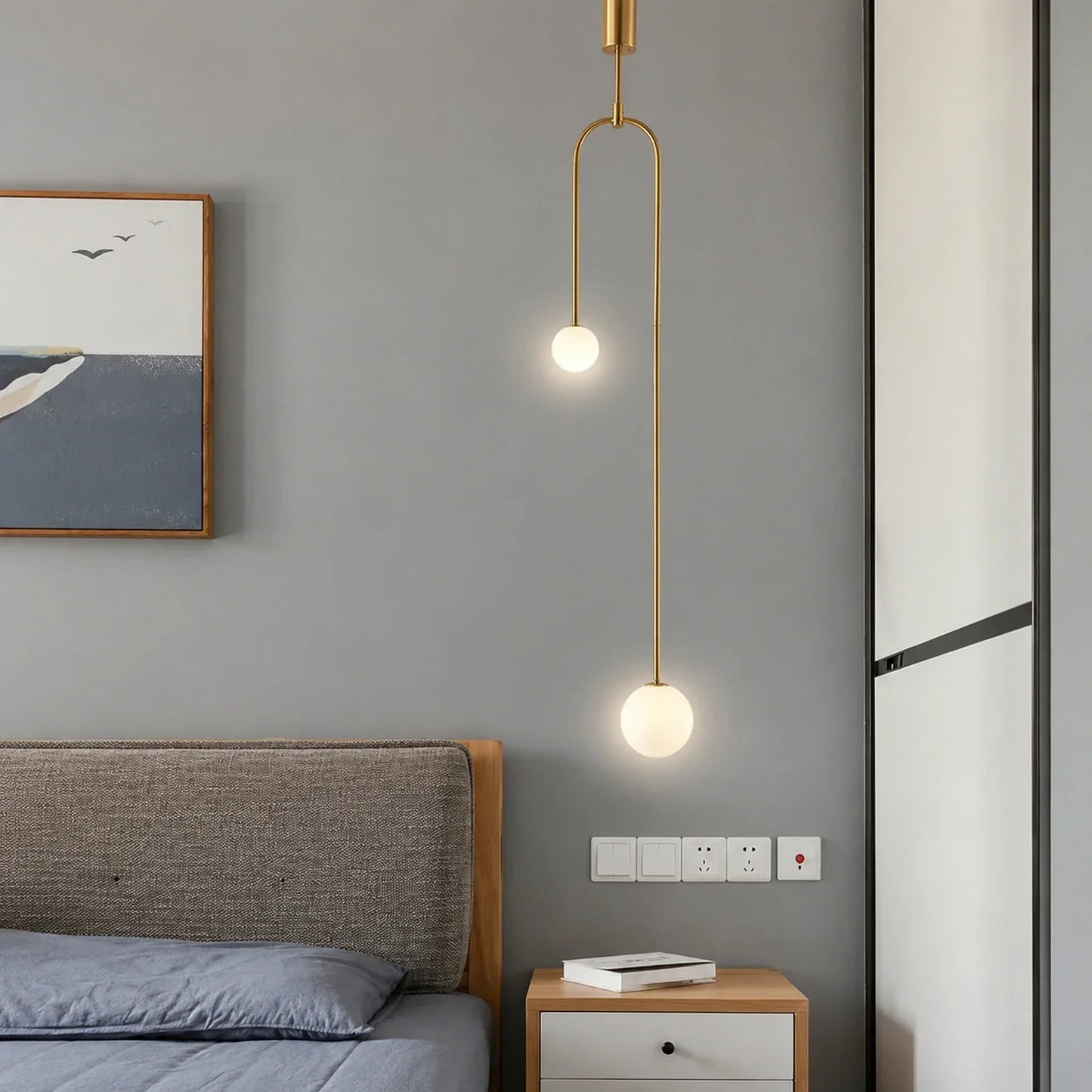 Modern double globe pendant light with a gold finish, suspended above a wooden bedside table in a minimalist bedroom with grey walls and wall art.