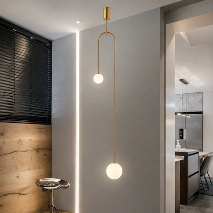 Modern double globe pendant light with a gold finish, featuring two illuminated white glass spheres suspended at different heights in a minimalist room.