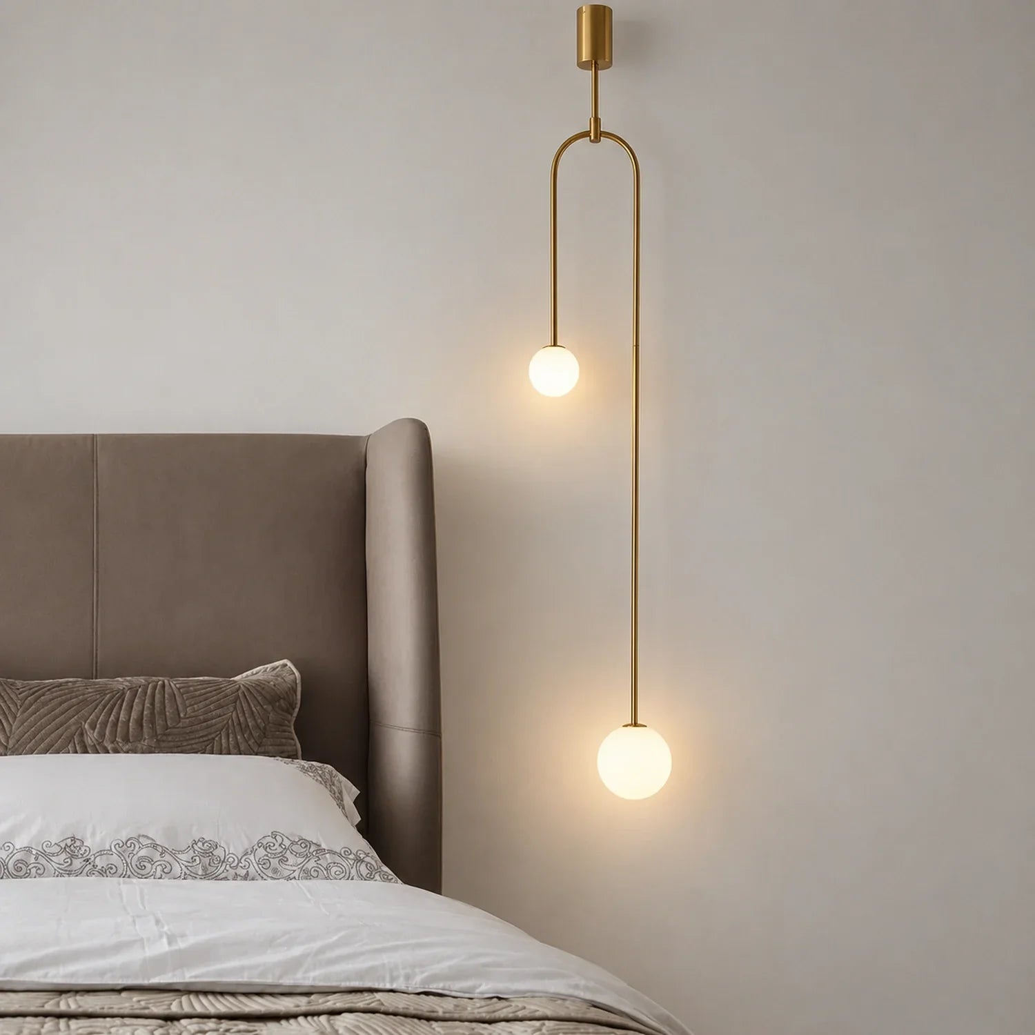 Modern double globe pendant light with a gold finish hanging beside a neutral upholstered bed in a minimalist bedroom setting.
