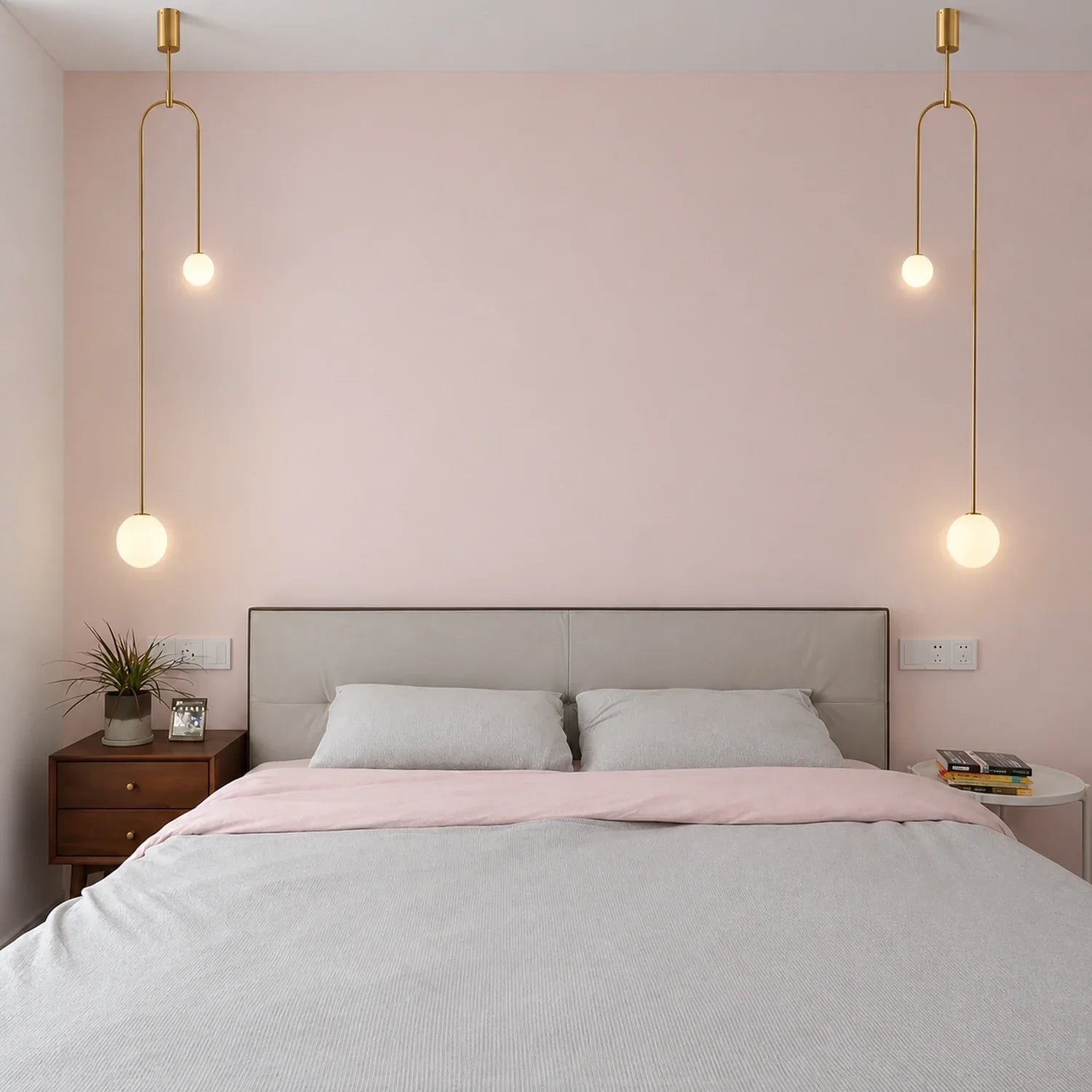 Two modern double globe pendant lights with brass fixtures hanging symmetrically above a minimalist bedroom headboard against a soft pink wall.