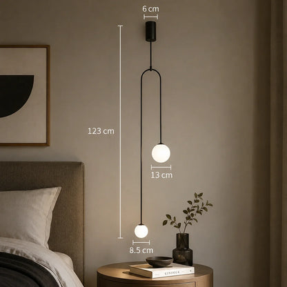 Modern black double globe pendant light with dimensions shown, featuring two white glass spheres suspended at different heights above a bedside table.