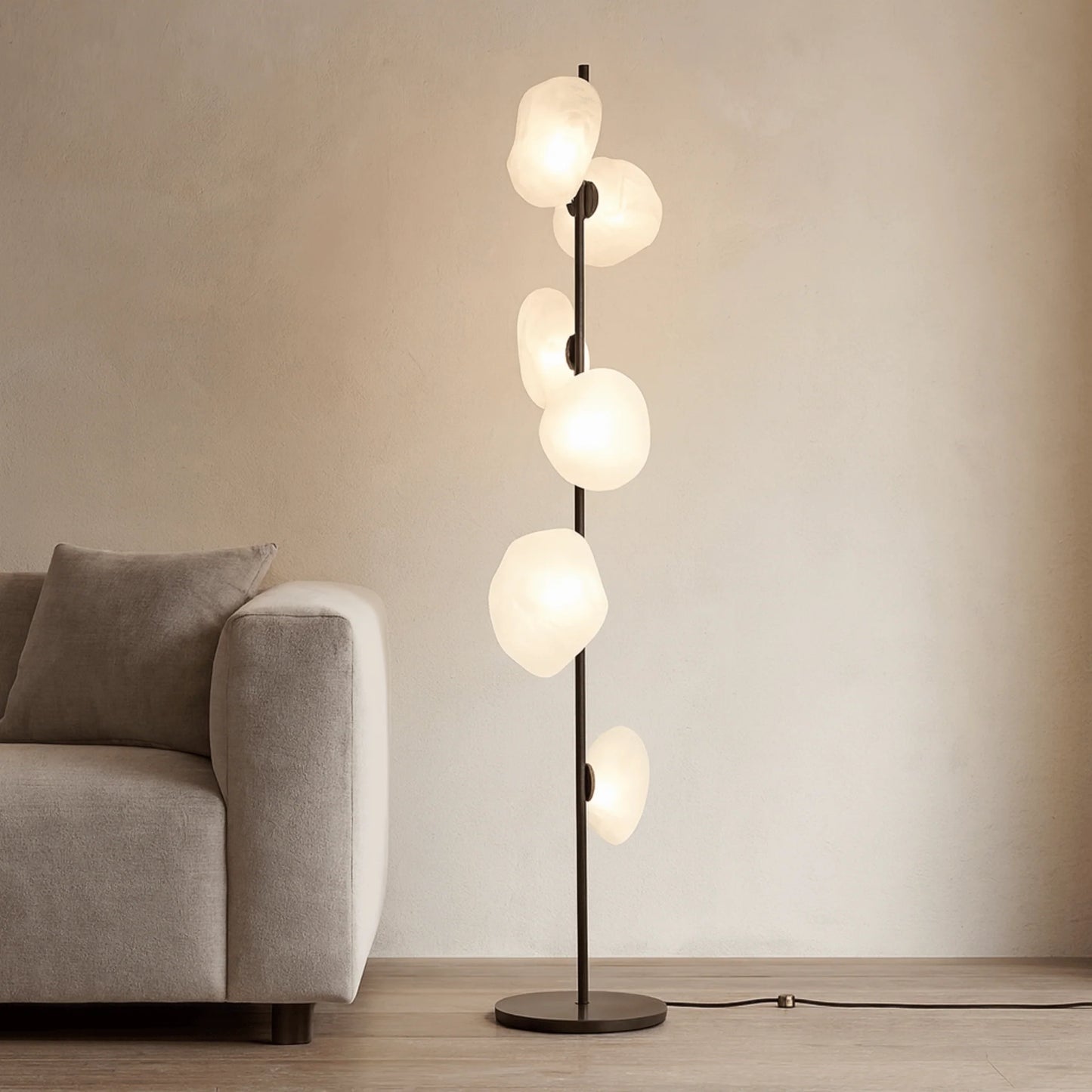 Modern black LED floor lamp with six glowing, organic-shaped white glass shades, positioned next to a grey fabric sofa in a minimalist living room.