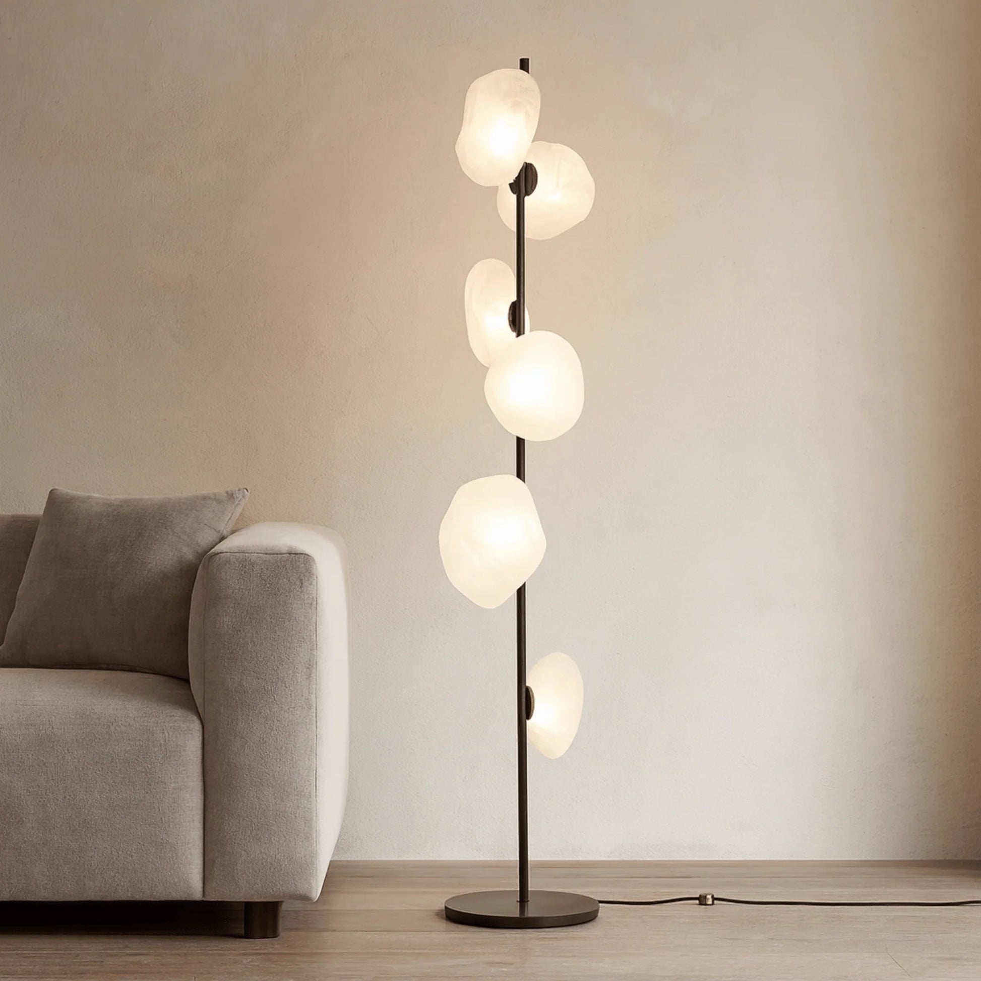 Modern black LED floor lamp with six glowing, organic-shaped white glass shades, positioned next to a grey fabric sofa in a minimalist living room.