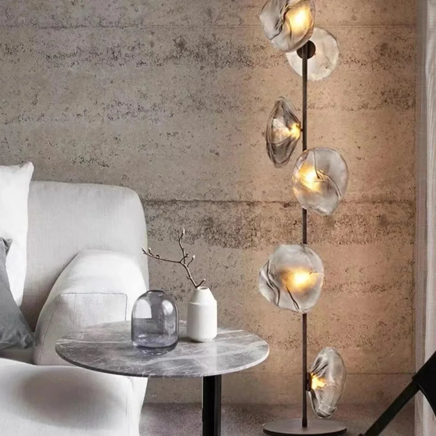 A modern glass LED floor lamp with organic, textured grey glass shades glowing with warm light next to a marble side table and grey armchair.