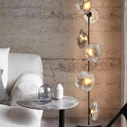 A modern glass LED floor lamp with organic, textured grey glass shades glowing with warm light next to a marble side table and grey armchair.