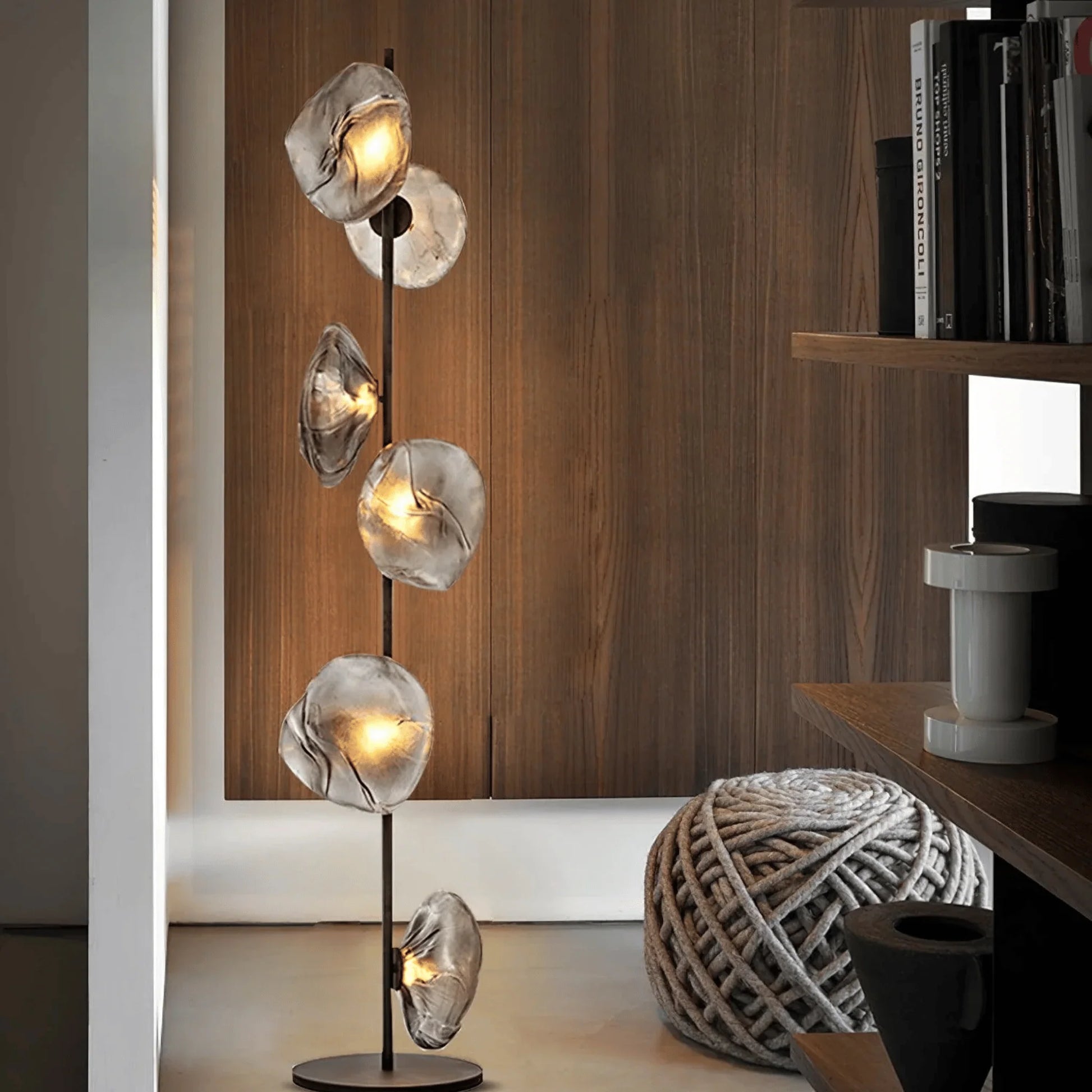 Modern glass LED floor lamp featuring six textured, organic-shaped glass shades glowing warmly next to a wooden wall and grey rope pouf.