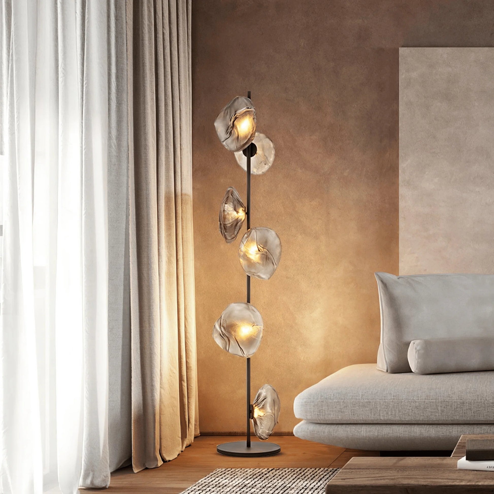 Modern glass LED floor lamp with six glowing, organic-shaped glass shades on a black stem, positioned next to a grey sofa in a warm living room.