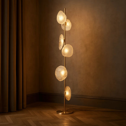 Modern gold LED floor lamp with six organic-shaped transparent glass shades emitting a warm glow, standing on a parquet floor in a cosy room.